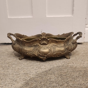 May include: An elongated, oval-shaped brass bowl featuring ornate, raised relief detailing, a scalloped rim, and two side handles. The vessel is supported by four small, decorative feet and displays a muted, metallic gold finish with intricate patterns.