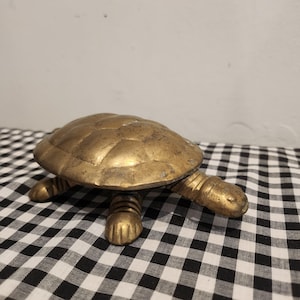May include: A brass turtle figurine with a polished, metallic gold finish. The object features a rounded, segmented shell and four legs with visible claw details. The head is extended forward, and the surface shows natural patina and texture.