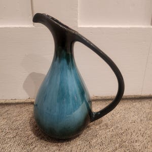 May include: A ceramic pitcher with a teardrop shape and a curved handle. The top and handle are black, fading into teal and blue tones on the body. The pitcher has a long spout and is likely used for serving drinks.