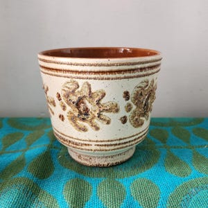 May include: Cream ceramic plant pot with brown floral patterns and a brown rim. The pot has a textured surface and a pedestal base. The floral design is repeated around the body of the pot.