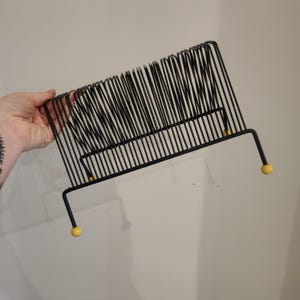 May include: Black metal record holder with a wire design. The holder has a rectangular shape with vertical wires and yellow ball feet. The holder is designed to hold vinyl records and is a vintage style.
