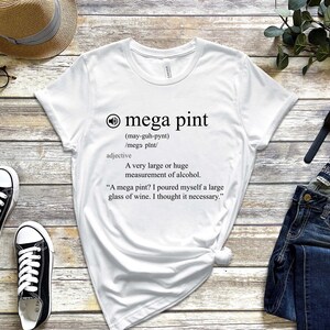 以下が含まれることがあります： 「メガパイント」という言葉の定義が書かれた白いTシャツ。メガパイントは、非常に大きく、または巨大なアルコールの量を指す言葉です。テキストには、単語の発音と、その使用例がユーモラスに書かれています。