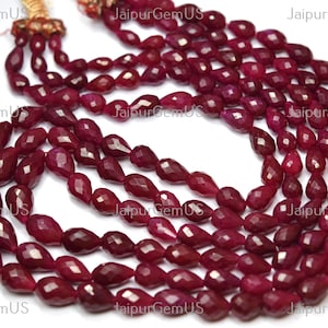 May include: A multi-strand necklace featuring faceted, teardrop-shaped ruby beads. The deep red gemstones are strung together, creating a layered effect. The necklace has a gold-colored clasp and is displayed against a white background.
