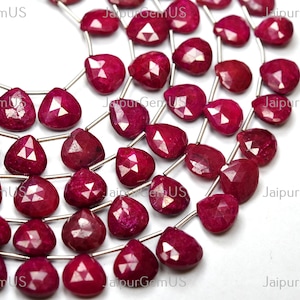 May include: Strands of faceted, heart-shaped ruby gemstones. The deep red stones are strung on silver-colored wire, with multiple strands visible. The gemstones are arranged against a white background, with the text "JaipurGemUS" visible.