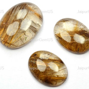 May include: Three oval-shaped gemstones with a clear, golden-brown hue. The stones feature intricate, needle-like inclusions, creating a striking visual texture. The gemstones are displayed against a white background. The text "JaipurGemUS" is visible.