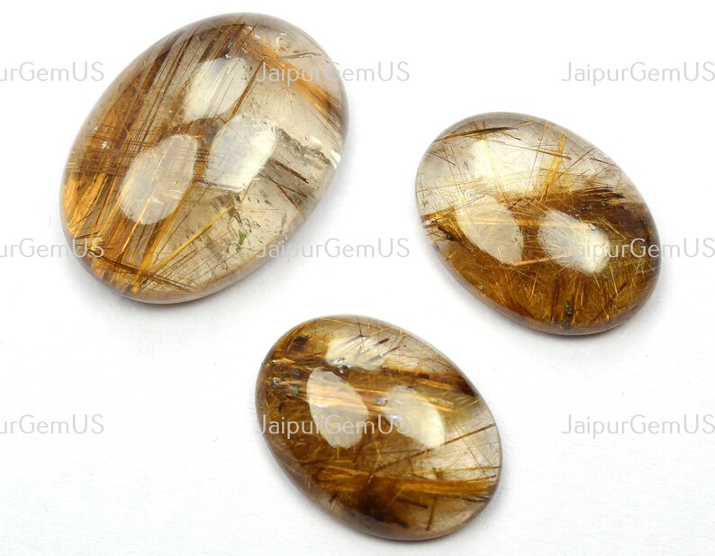 May include: Three oval-shaped gemstones with a clear, golden-brown hue. The stones feature intricate, needle-like inclusions, creating a striking visual texture. The gemstones are displayed against a white background. The text "JaipurGemUS" is visible.