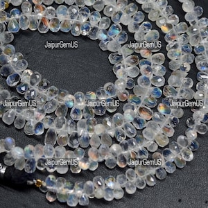 May include: Strands of clear, faceted gemstone beads with iridescent flashes of color. The beads are teardrop-shaped and strung together, creating a necklace or bracelet. The text "JaipurGemUS" is visible in several places.