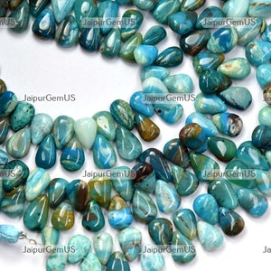 Op de afbeelding: Een verzameling gepolijste, druppelvormige edelstenen in tinten teal, turquoise en bruin. De stenen zijn in een los patroon gerangschikt, met een kwast zichtbaar aan de linkerkant. De edelstenen zijn waarschijnlijk voor het maken van sieraden.