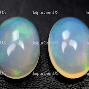 May include: Two oval opals with a polished, iridescent surface. The opals display a play of colour, with flashes of blue, green, and yellow. The gemstones are set against a dark background, highlighting their luminous quality.