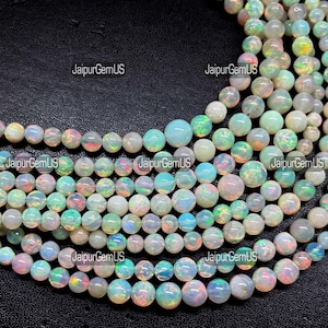 Peut inclure: Une collection de perles d'opale rondes et translucides disposées en plusieurs rangs. Les perles présentent une couleur de base blanc laiteux avec des éclats irisés de lumière verte, bleue et rose. Le texte JaipurGemUS est répété sur l'image.