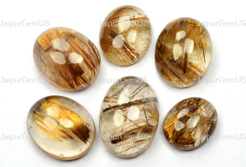 May include: Six oval-shaped gemstones with a golden-brown, hair-like inclusions. The stones are translucent with a polished surface, set against a white background. The text "Jaipur Gem US" is visible.