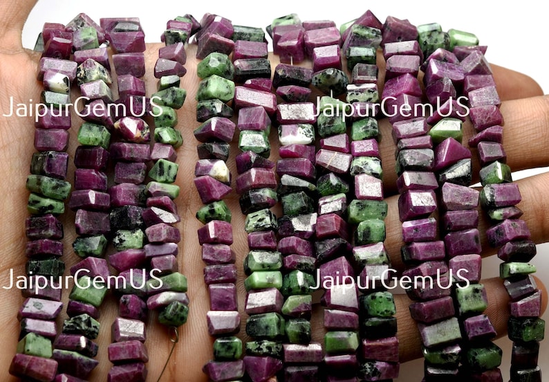 Puede incluir: Hebras de cuentas de piedras preciosas de rub&iacute; y zoisita en bruto. Las cuentas son una mezcla de colores rojo intenso y verde, con un corte rugoso e irregular. Las cuentas est&aacute;n ensartadas, listas para la fabricaci&oacute;n de joyas. El texto "JaipurGemUS" es visible.