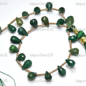 Op de afbeelding: Strengen gefacetteerde, smaragdgroene edelsteen kralen. De kralen zijn druppel- en rond van vorm, geregen op goudkleurige draad. Groene kwasten met gouden accenten zijn zichtbaar aan de uiteinden. De afbeelding heeft de tekst "JaipurGemUS".