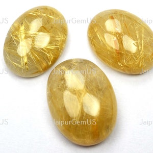 May include: Three oval-shaped golden rutilated quartz gemstones. The stones have a translucent, honey-colored base with striking golden needle-like inclusions. The gemstones are set against a white background.