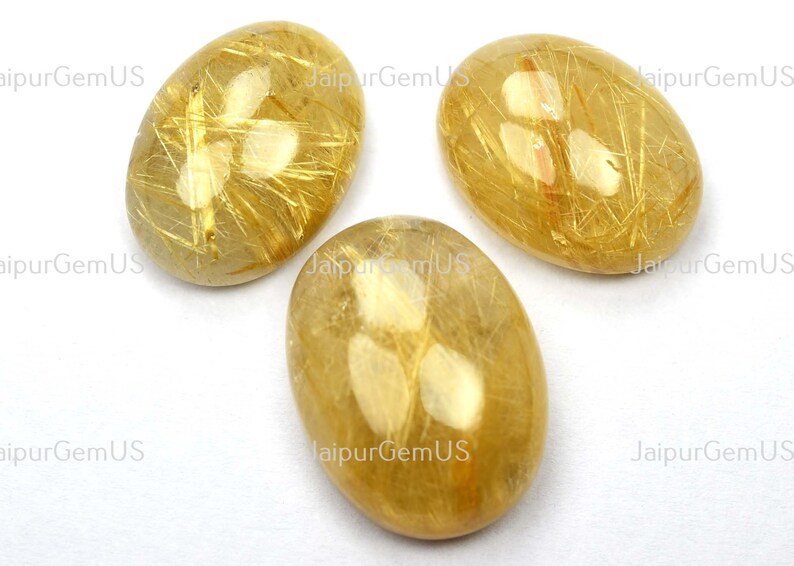 May include: Three oval-shaped golden rutilated quartz gemstones. The stones have a translucent, honey-colored base with striking golden needle-like inclusions. The gemstones are set against a white background.