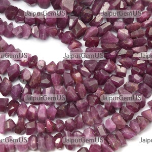May include: A collection of raw ruby gemstones, each cut with multiple facets. The deep red-purple stones are strung together, creating a textured display. The gemstones are displayed against a white background. Each stone is approximately 0.5-1 cm.