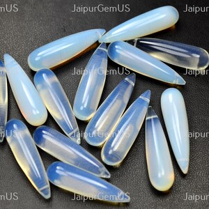 May include: A collection of teardrop-shaped opalescent gemstones. The stones are a translucent white with a subtle blue hue, and are arranged on a dark surface. The gemstones are elongated, with a pointed tip and a rounded base.