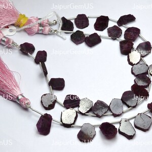 May include: Strands of raw, dark red ruby gemstone beads. The beads are strung on silver wire and are irregularly shaped. Pink tassels with silver accents are visible in the image. The text "Jaipur Gem US" is visible in the image.
