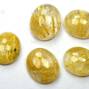 May include: Five oval-shaped, polished gemstones. The stones are a translucent golden color with internal golden needle-like inclusions. The gemstones are displayed on a white surface.