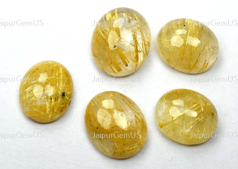 May include: Five oval-shaped, polished gemstones. The stones are a translucent golden color with internal golden needle-like inclusions. The gemstones are displayed on a white surface.