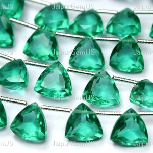 Könnte beinhalten: Nahaufnahme von mehreren Strängen facettierter, dreieckiger, smaragdgrüner Edelsteine. Jeder Edelstein ist auf einem silberfarbenen Draht aufgefädelt. Die Edelsteine sind durchscheinend und reflektieren das Licht, wodurch ein funkelnder Effekt entsteht. Der Hintergrund ist weiß.