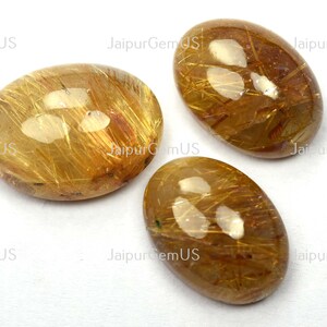 May include: Three oval-shaped gemstones with a golden-brown hue and internal golden needle-like inclusions. The stones are polished and have a glossy surface, set against a white background. The text "JaipurGemUS" is visible.