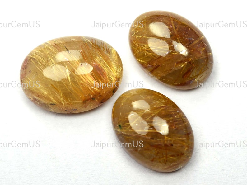 May include: Three oval-shaped gemstones with a golden-brown hue and internal golden needle-like inclusions. The stones are polished and have a glossy surface, set against a white background. The text "JaipurGemUS" is visible.