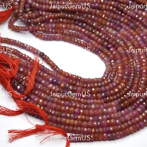 May include: Strands of faceted ruby gemstone beads in varying shades of red and pink. The beads are strung together, with red tassels visible on the left side. The image is labeled with "Jaipur GemUS".