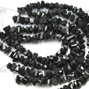 May include: Strands of black, irregularly shaped gemstone beads. The beads are strung together, creating a flowing, organic pattern. The gemstones have a polished, reflective surface, and are likely used for jewelry making.