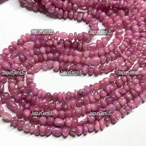Puede incluir: Primer plano de hilos de piedras preciosas rosas pulidas. Las cuentas son pequeñas, de forma irregular y pulidas. Las piedras preciosas son de un color rosa intenso y rosado. La imagen está bien iluminada, mostrando la textura y el color de las piedras.