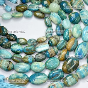 Op de afbeelding: Strengen ovale edelsteen kralen in tinten blauw, turquoise en bruin. De kralen zijn aan elkaar geregen, met een kwast aan het einde van elke streng. De kralen hebben een gepolijst oppervlak. De tekst "JaipurGemUS" is inbegrepen.