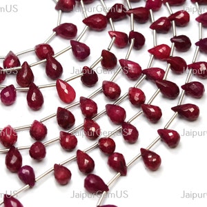 May include: Strands of faceted, teardrop-shaped ruby gemstones in varying sizes. The deep red stones are strung on thin silver wires, creating a shimmering effect. The gemstones are displayed against a white background.