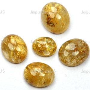 May include: Five oval-shaped golden rutilated quartz gemstones. The stones have a translucent appearance with golden needle-like inclusions. The gemstones are set against a white background, with the text "JaipurGemUS" visible.