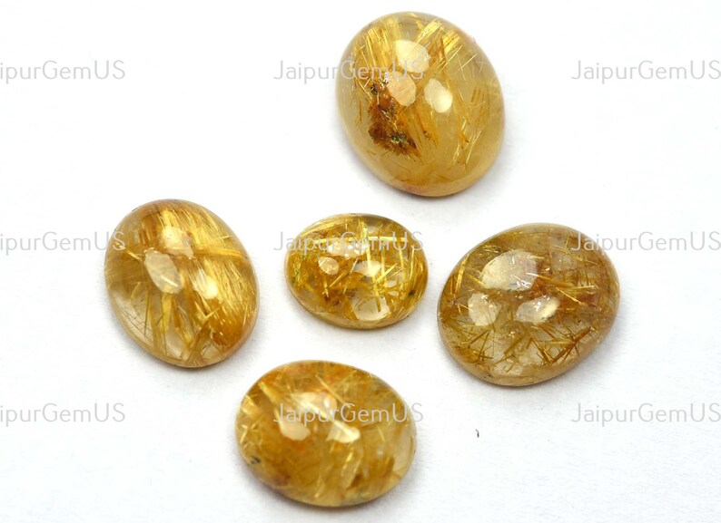 May include: Five oval-shaped golden rutilated quartz gemstones. The stones have a translucent appearance with golden needle-like inclusions. The gemstones are set against a white background, with the text "JaipurGemUS" visible.