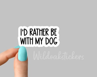 Op de afbeelding: Witte sticker met zwarte tekst die "Ik zou liever bij mijn hond zijn" zegt. De sticker wordt vastgehouden door een hand met lichtblauwe nagellak.