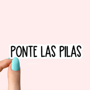 Könnte beinhalten: Ein weißer Aufkleber mit schwarzem Text, der "PONTE LAS PILAS" lautet, der von einer Hand mit hellblauem Nagellack gehalten wird.