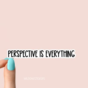 May include: A white sticker with black text that reads "Perspective is Everything". The sticker is being held by a hand with light blue nail polish.