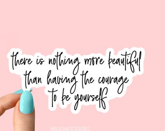 May include: White sticker with black cursive text that reads "There is nothing more beautiful than having the courage to be yourself." The sticker is being held by a finger with light blue nail polish.