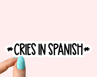 May include: White sticker with black text that reads "*CRIES IN SPANISH*" being held by a hand with light blue nail polish.