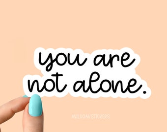 May include: White sticker with black text that reads "you are not alone." The sticker is being held by a hand with light blue nail polish.
