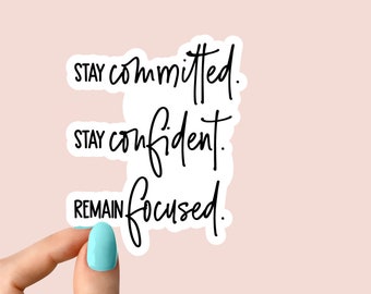 May include: A white sticker with black script that reads "Stay committed. Stay confident. Remain focused." The sticker is held by a hand with a light blue painted nail. The background is a light pink color.