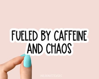 May include: A white sticker with the text "FUELED BY CAFFEINE AND CHAOS" in black, held by a hand with a light blue painted nail. The sticker is on a light pink background. The brand name "WILDOAKSTICKERS" is at the bottom.