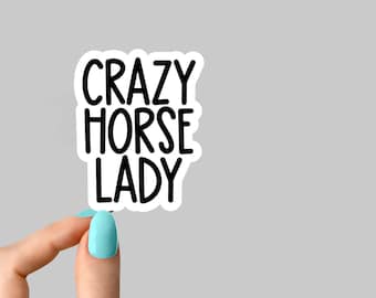 以下が含まれることがあります： 明るい青色のマニキュアを塗った手で、黒い文字で「CRAZY HORSE LADY」と書かれた白いステッカーを持っている。