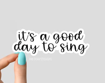 May include: A white sticker with the phrase "it's a good day to sing" in black cursive font. The sticker is held by a hand with a light blue painted fingernail. The text "WILDOAKSTICKERS" is at the bottom.