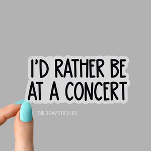 May include: A white sticker with the text "I'D RATHER BE AT A CONCERT" in black, sans-serif font. The sticker is held by a hand with a light blue nail polish. The sticker also has the text "WILDOAKSTICKERS" in a smaller font.