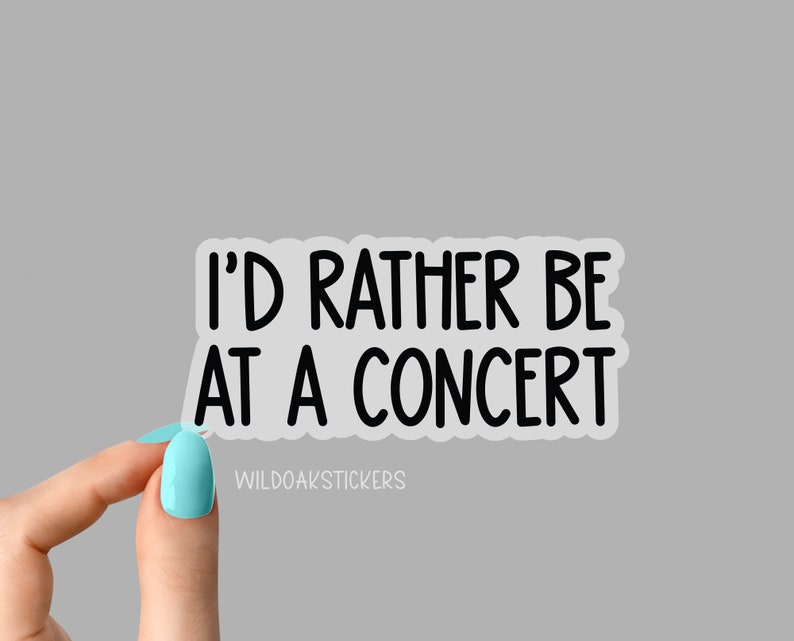 May include: A white sticker with the text "I'D RATHER BE AT A CONCERT" in black, sans-serif font. The sticker is held by a hand with a light blue nail polish. The sticker also has the text "WILDOAKSTICKERS" in a smaller font.
