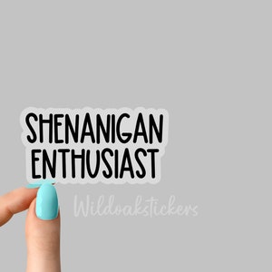 K&ouml;nnte beinhalten: Ein transparenter Aufkleber mit schwarzem Text, der "Shenanigan Enthusiast" lautet. Der Aufkleber wird von einer Hand mit hellblauem Nagellack gehalten.