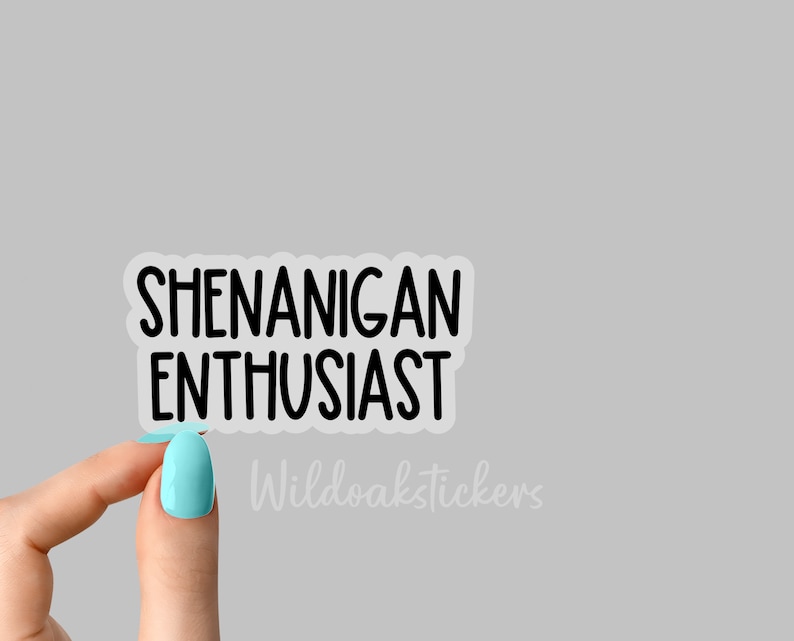 K&ouml;nnte beinhalten: Ein transparenter Aufkleber mit schwarzem Text, der "Shenanigan Enthusiast" lautet. Der Aufkleber wird von einer Hand mit hellblauem Nagellack gehalten.