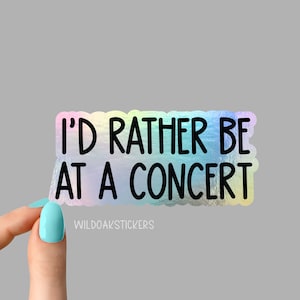 May include: A holographic sticker with the text "I'D RATHER BE AT A CONCERT" in black. The sticker has a scalloped edge and a rainbow sheen. The brand name "WILDOAKSTICKERS" is printed at the bottom.