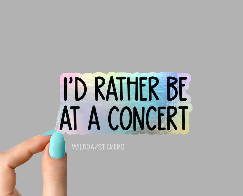 May include: A holographic sticker with the text "I'D RATHER BE AT A CONCERT" in black. The sticker has a scalloped edge and a rainbow sheen. The brand name "WILDOAKSTICKERS" is printed at the bottom.